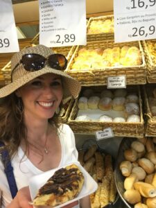 A smiling woman holds a cinnamon roll in front of a bakery display.