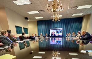 participants in a meeting sit around a board table with a video conference screen at one end. 