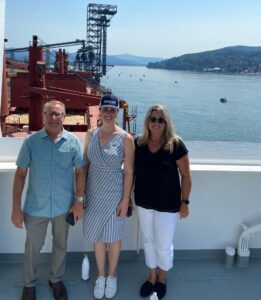 Three people stand on the deck of a ship with a river port visible in the background. 