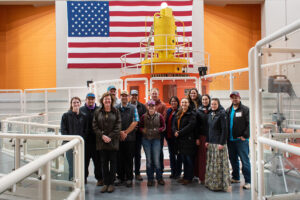 A group of people pose in front of a hydroelectric generator and American flag. 