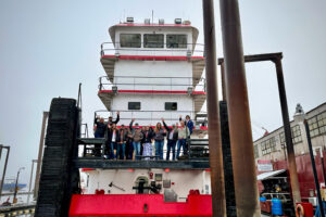 Group of people wave to the camera from the mid deck of a tug boat at a dock. 